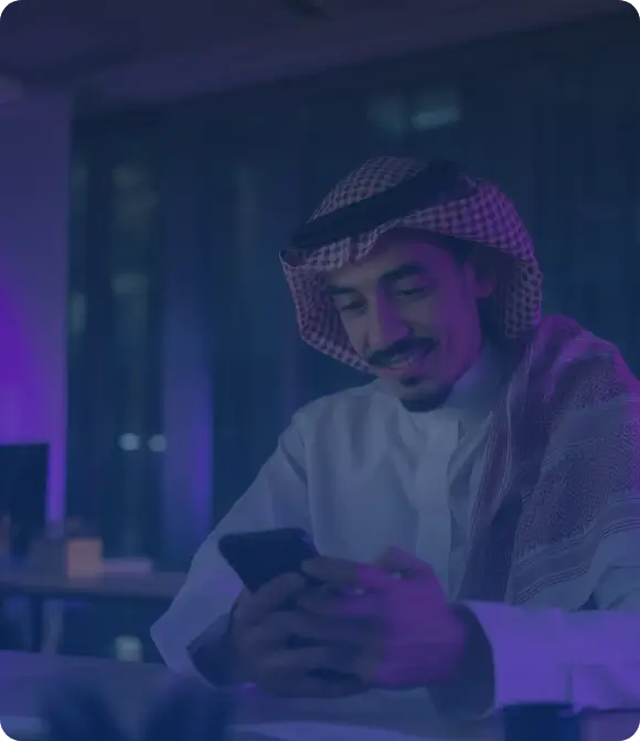 Person in traditional Middle Eastern attire sitting at a desk using a smartphone in a modern office with large windows and purple ambient lighting, conveying a professional and focused atmosphere