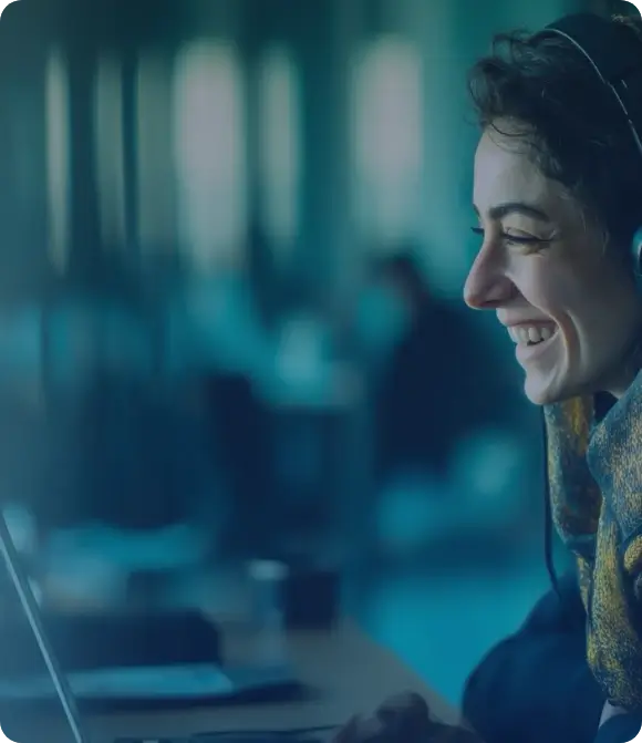Person wearing headphones sits at a desk working on a laptop in a modern office with a calm and focused atmosphere. The background is softly blurred, showing other people and desks, suggesting a collaborative and professional environment.