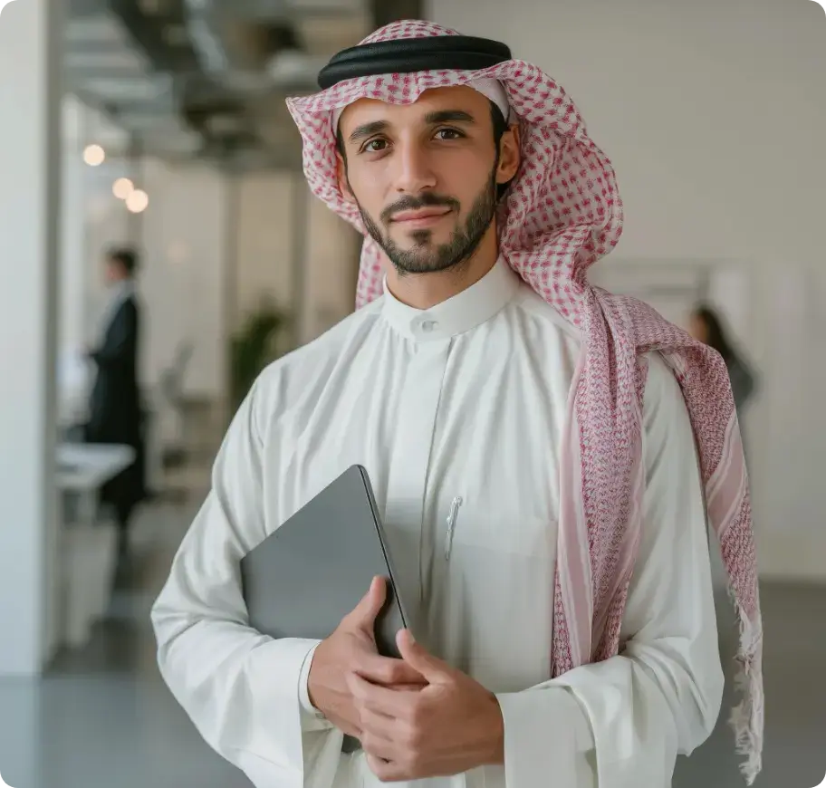 رجل يرتدي الزي الخليجي يحمل حاسوبًا مغلقًا في بيئة مكتبية عصرية وهادئة تدل على الثقة والاحتراف
